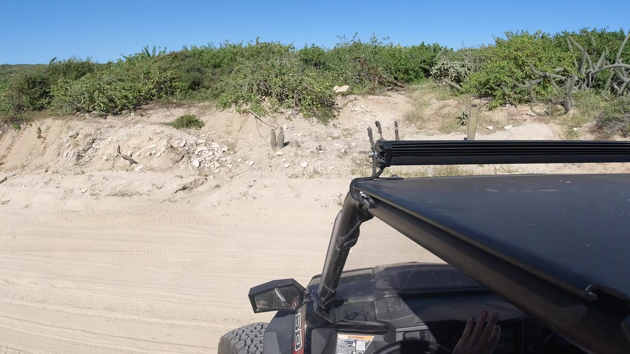 Opening view from a sand car in Los Cabos, Mexico, with a black roof frame in the foreground, pale sand road, green scrub, and no visible people.