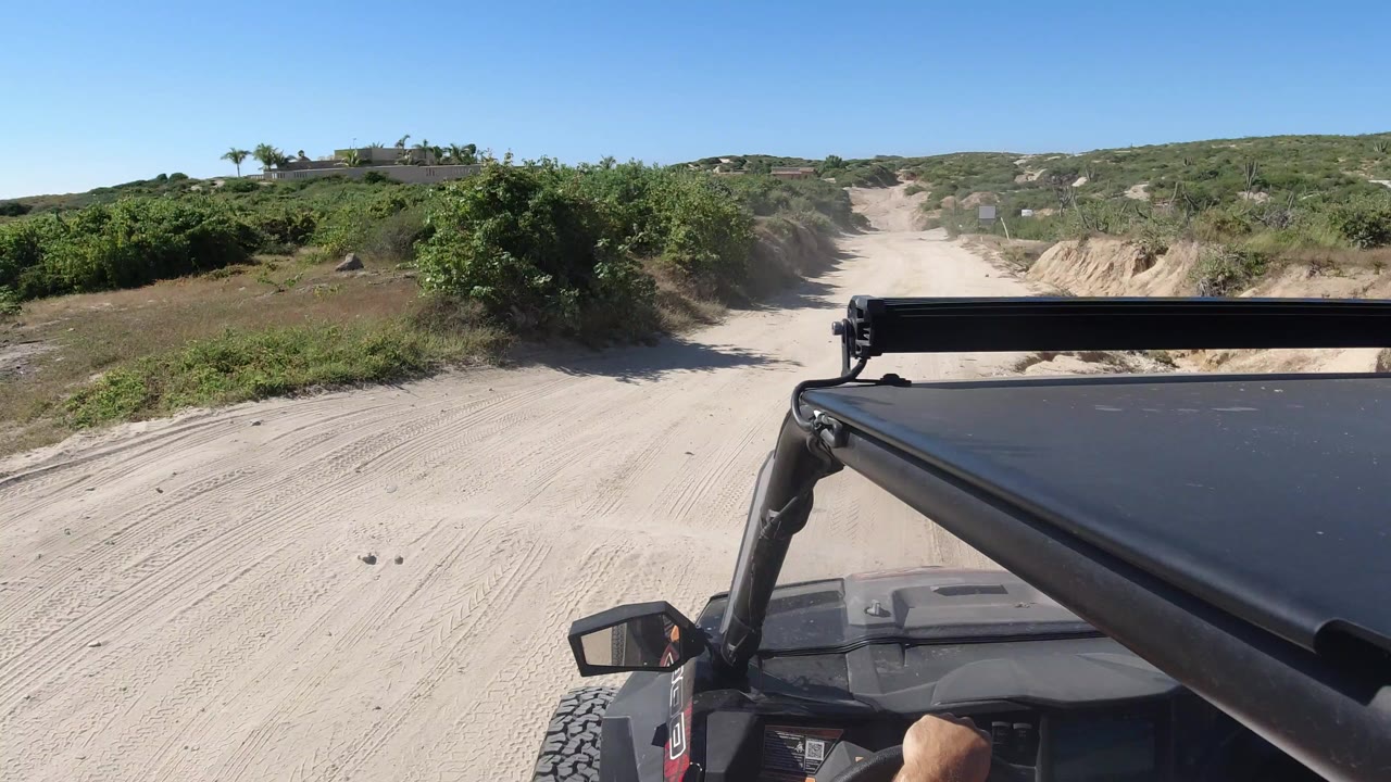 Opening view from an off-road vehicle on a sandy road in Los Cabos, Mexico, with green scrub on both sides, low hills ahead, and no visible people.