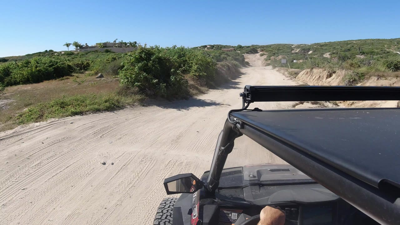 The vehicle continues toward a pale sandy track in Los Cabos, Mexico, with brushy banks, tire marks, a clear sky, and no visible faces.