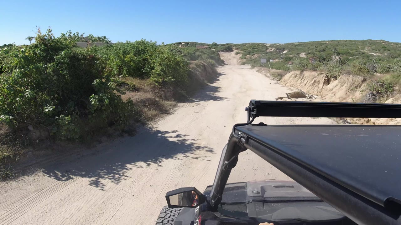 Sandy road curves past dense shrubs and a vehicle hood edge.
