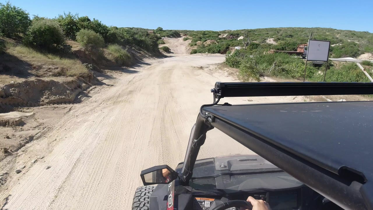 The off-road vehicle foreground remains close to camera as the sandy Los Cabos road rises toward low green hills, with a small sign and vegetation along the roadside.