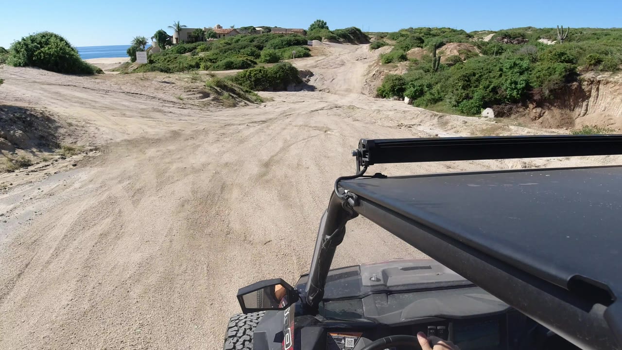 The buggy continues along a pale sand track in Los Cabos, Mexico, with the vehicle roof edge at lower right, low shrubs across the dunes, and blue water visible behind them.