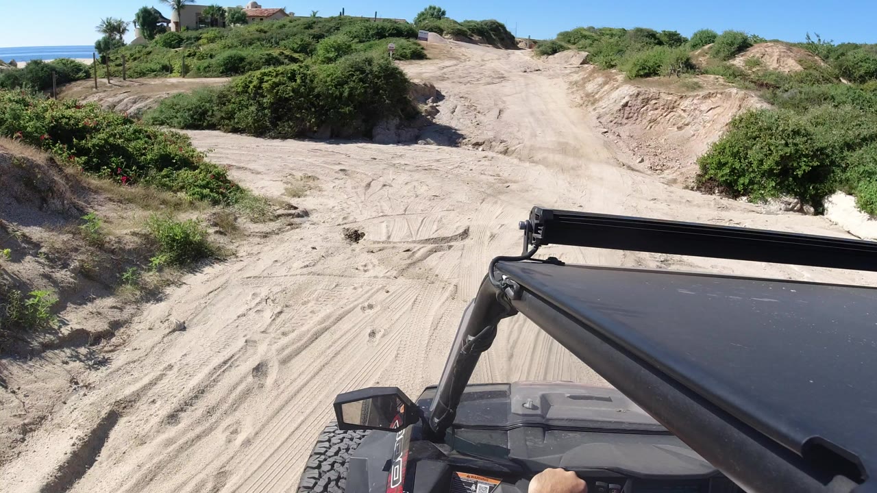 Forward view from a black off-road vehicle on a sandy Los Cabos road, with green bushes, eroded banks, blue sky, distant houses, and no visible faces.