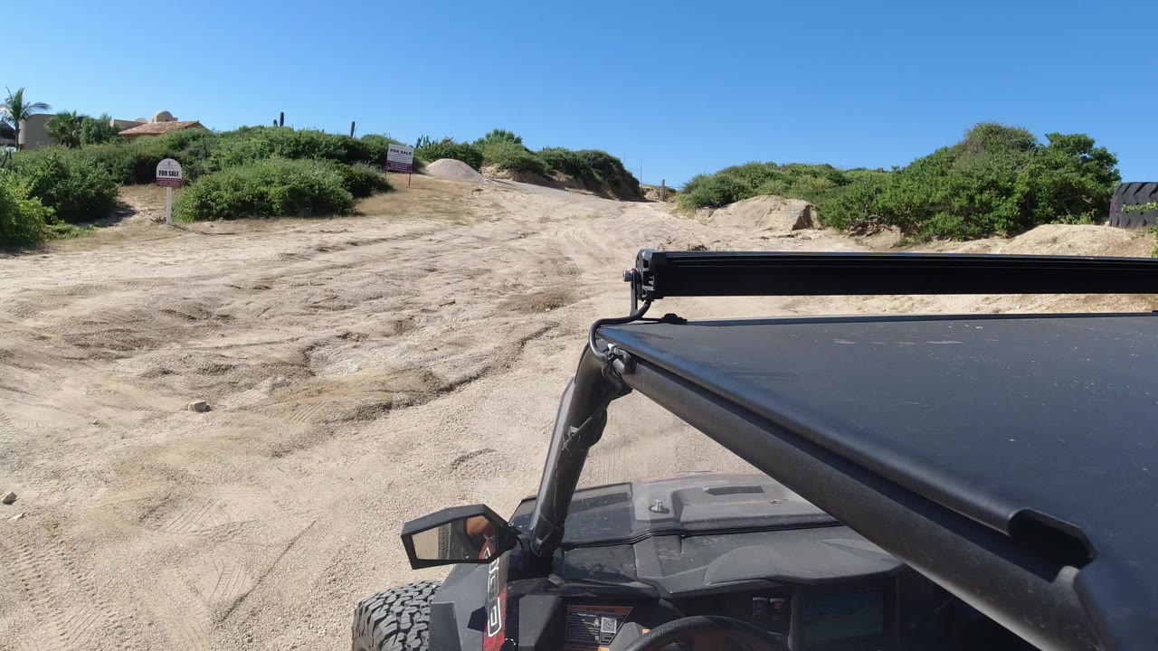 Passenger-side view over the black vehicle roof in Los Cabos, Mexico, looking toward a pale dirt road, dense green shrubs, small signs, and no visible faces.