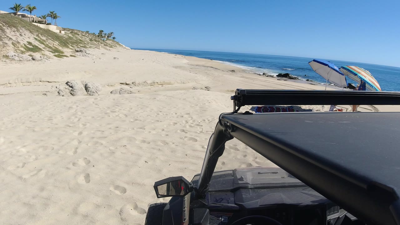 Parked dark car beside a sandy beach with a striped parasol and blue ocean.