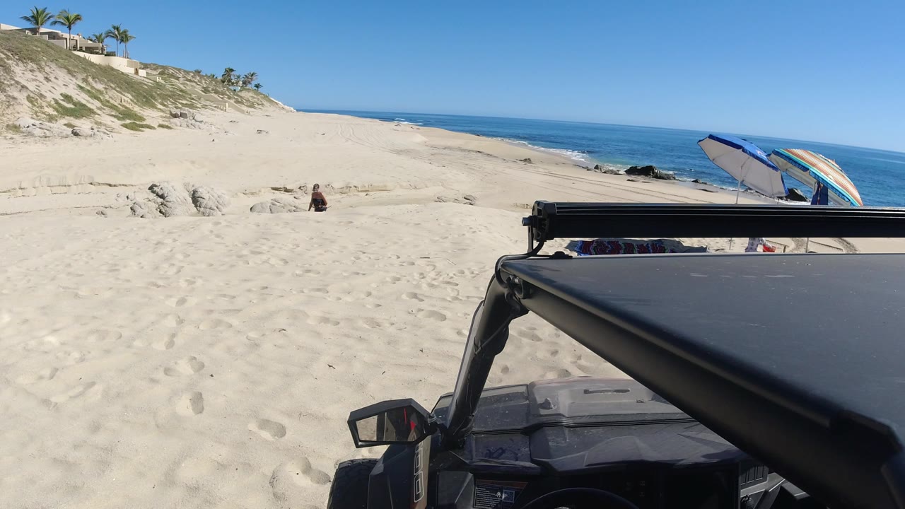 Parked vehicle beside sandy beach with ocean and umbrella beyond.
