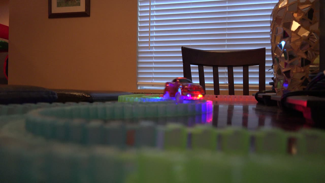 In Denver, Colorado, a low table-level view shows glowing toy train lights on a colorful track in front of a dining chair and closed window blinds, with no visible people.