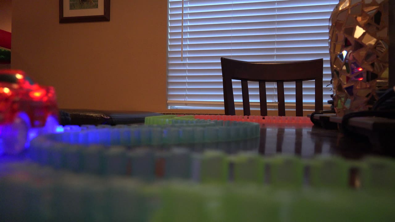 In Denver, Colorado, the toy train lights appear blurred near the left edge while the colorful track crosses the dining room table below a chair and closed blinds, with no visible people.