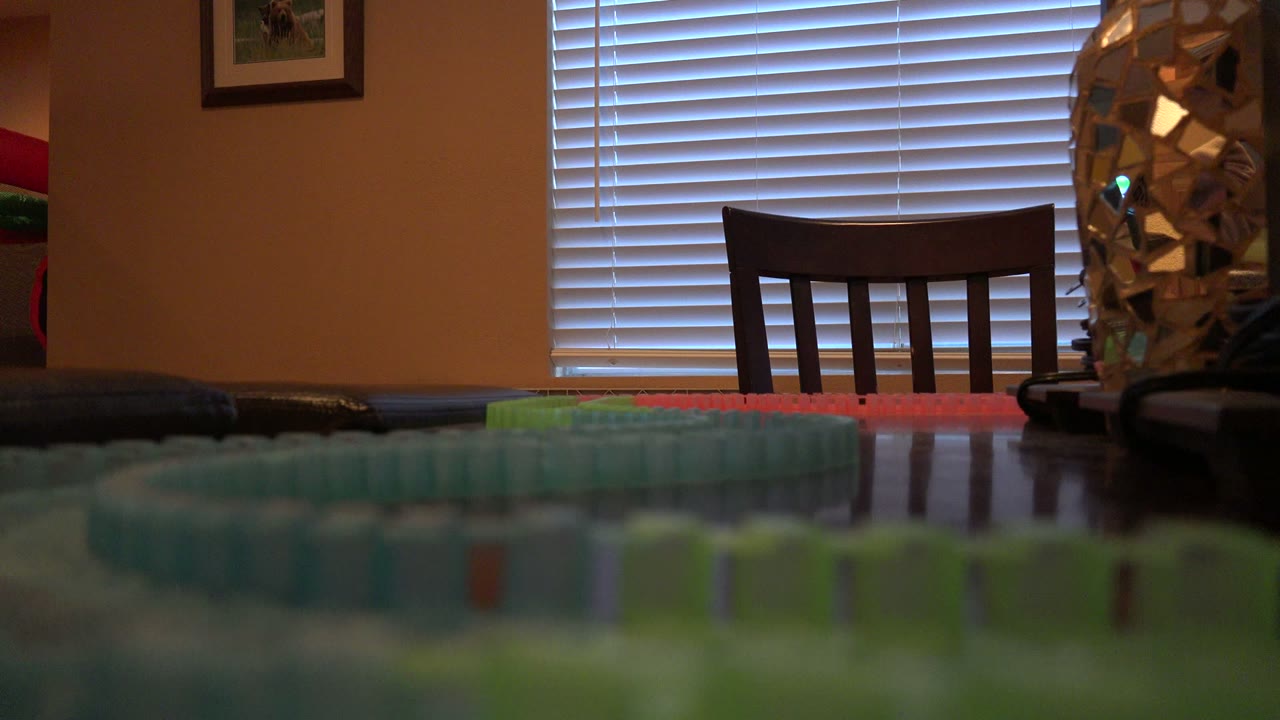 Denver 2016 frame showing the toy train track loop blurred along the lower foreground, with a chair centered against bright horizontal blinds and no visible people.