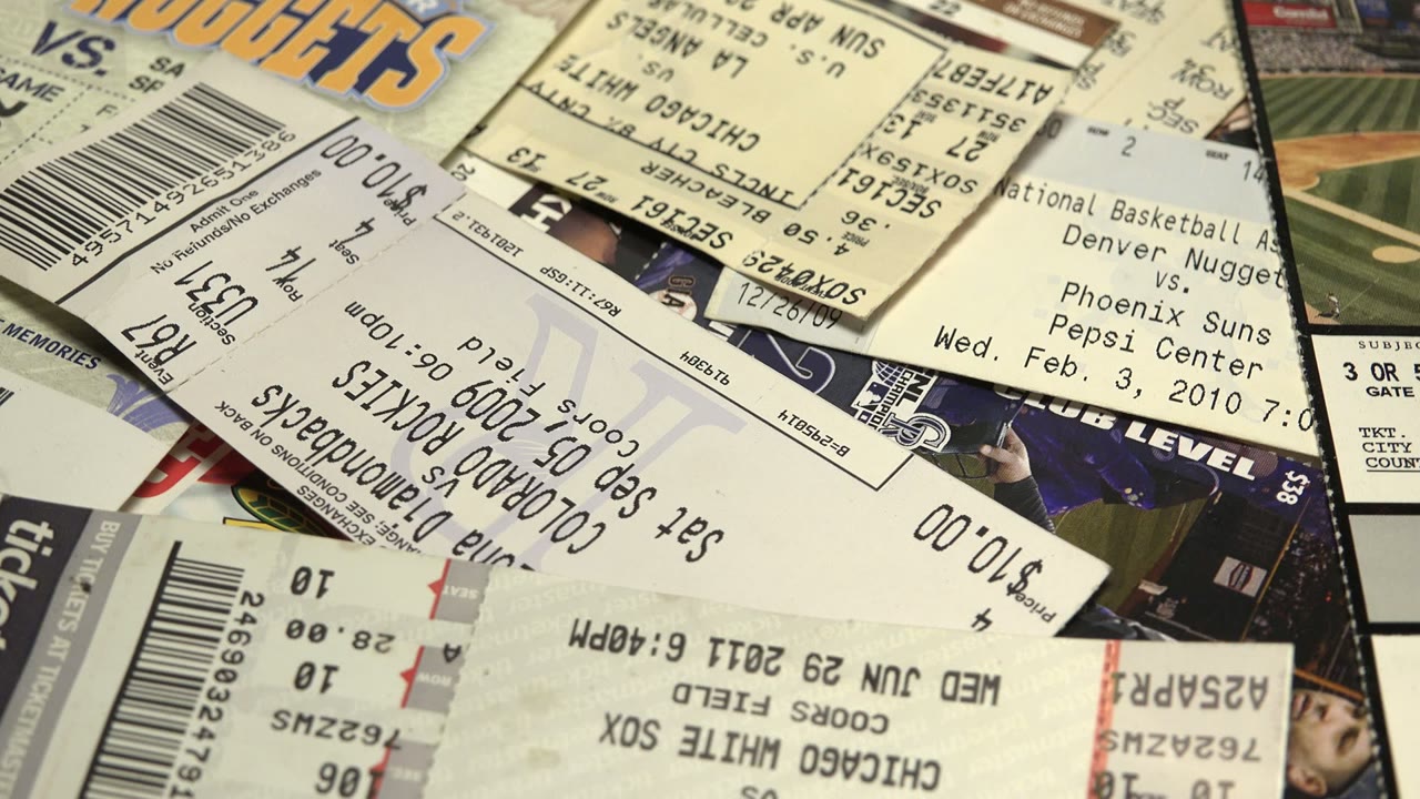 Layered paper tickets and receipts fill the frame, including visible price amounts, barcodes, basketball ticket text, and baseball ticket fragments, with no visible people.