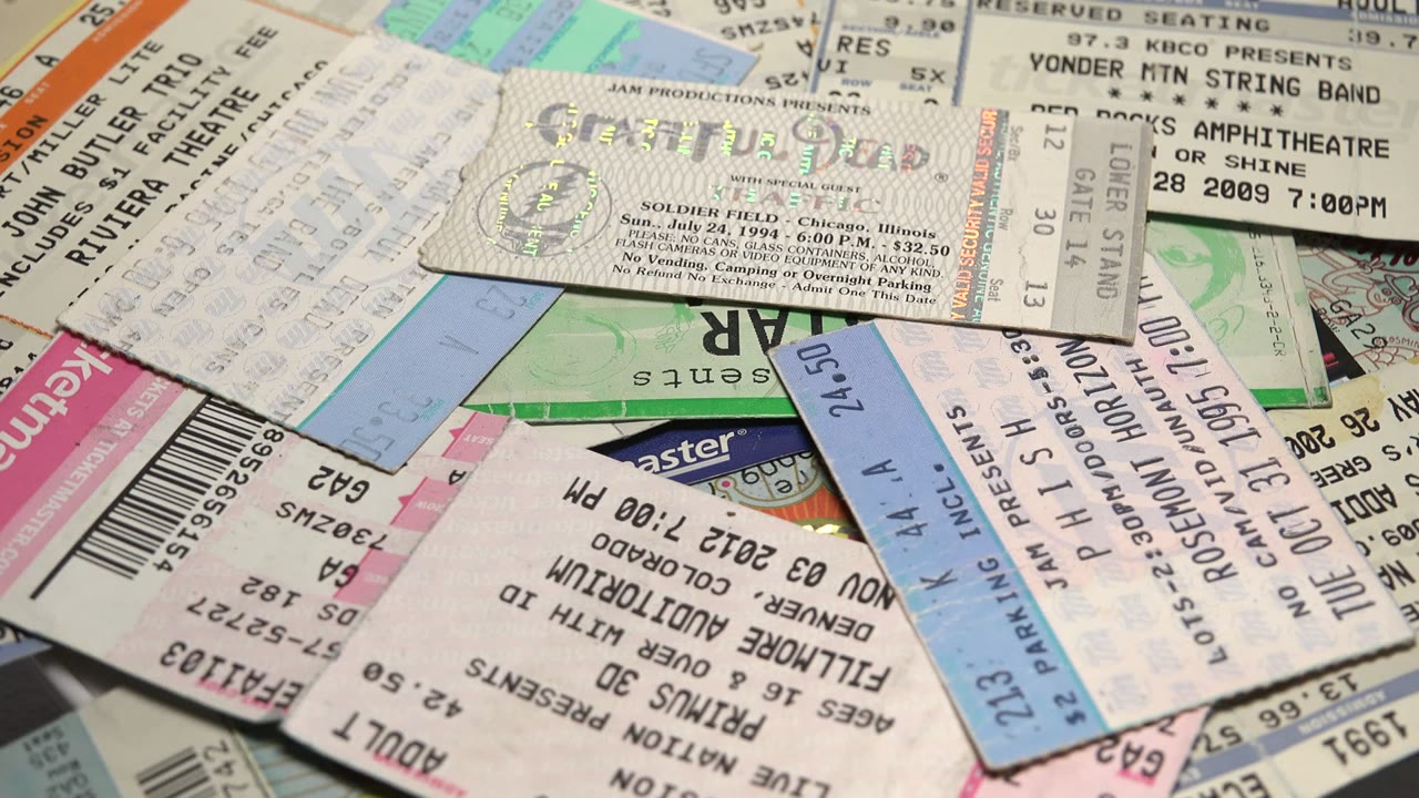 A dense close-up of overlapping concert tickets, event passes, barcodes, and pastel paper sections covering the table, with no visible people.