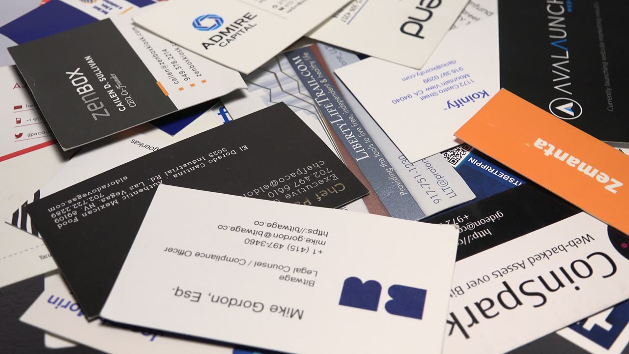 Close-up card pile with a white business card near center, black cards around it, and an orange promotional card angled across the upper right, with no visible people.