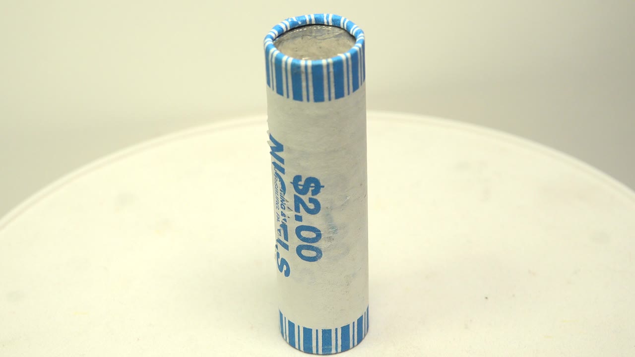 An upright paper-wrapped roll of nickels sits near the center of a white turntable, with blue wrapper text partly visible and no visible people.