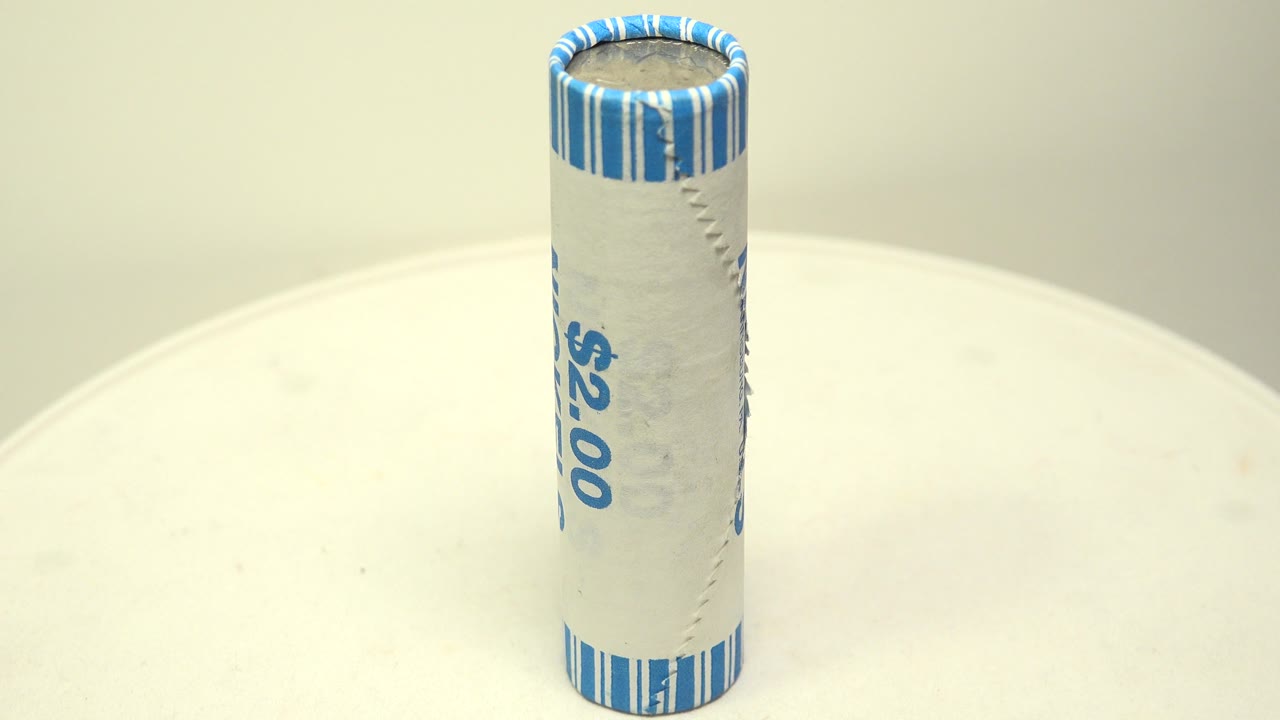 A two dollar paper roll of nickels stands upright near the center of a white surface, with blue striped crimping visible at the top and no visible people.