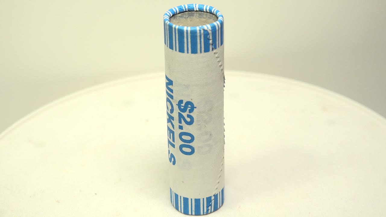 The upright coin roll has rotated so partial blue wrapper text appears along the side, while the top crimp and white tabletop stay sharply visible with no visible people.