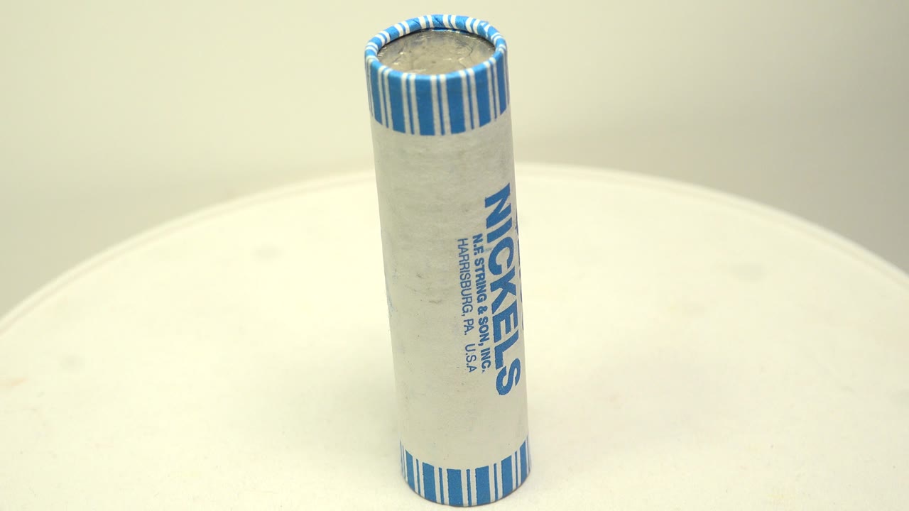 A paper-wrapped roll of nickels stands upright near the center of a white circular board, with blue striped wrapper ends and no visible people.