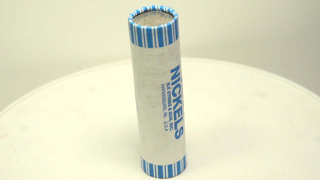 The coin roll remains upright on the white circular board, with the blue NICKELS label facing the camera more clearly and no visible people.