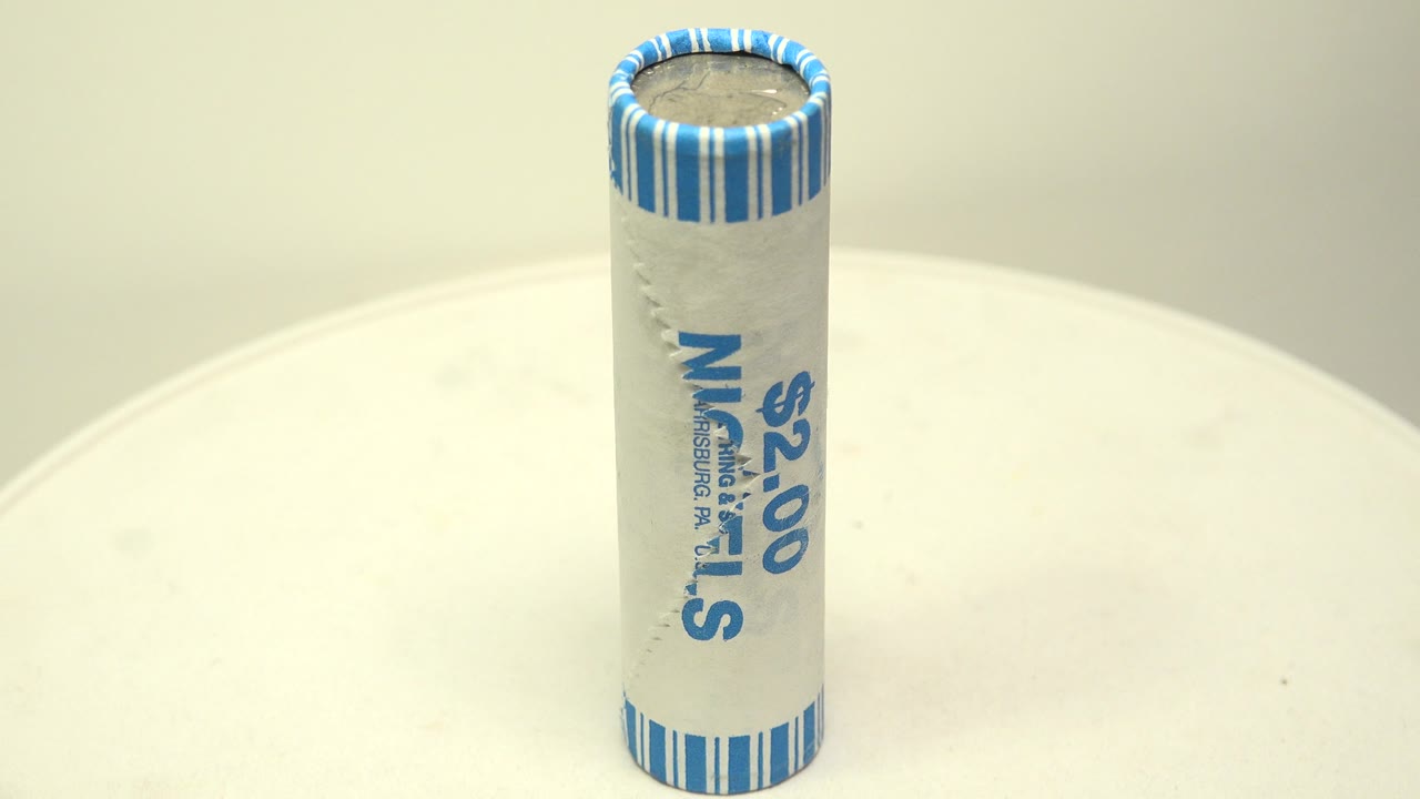 An upright paper-wrapped roll of nickels stands near center frame on a pale white surface, with blue striped paper visible at the top and no visible people.