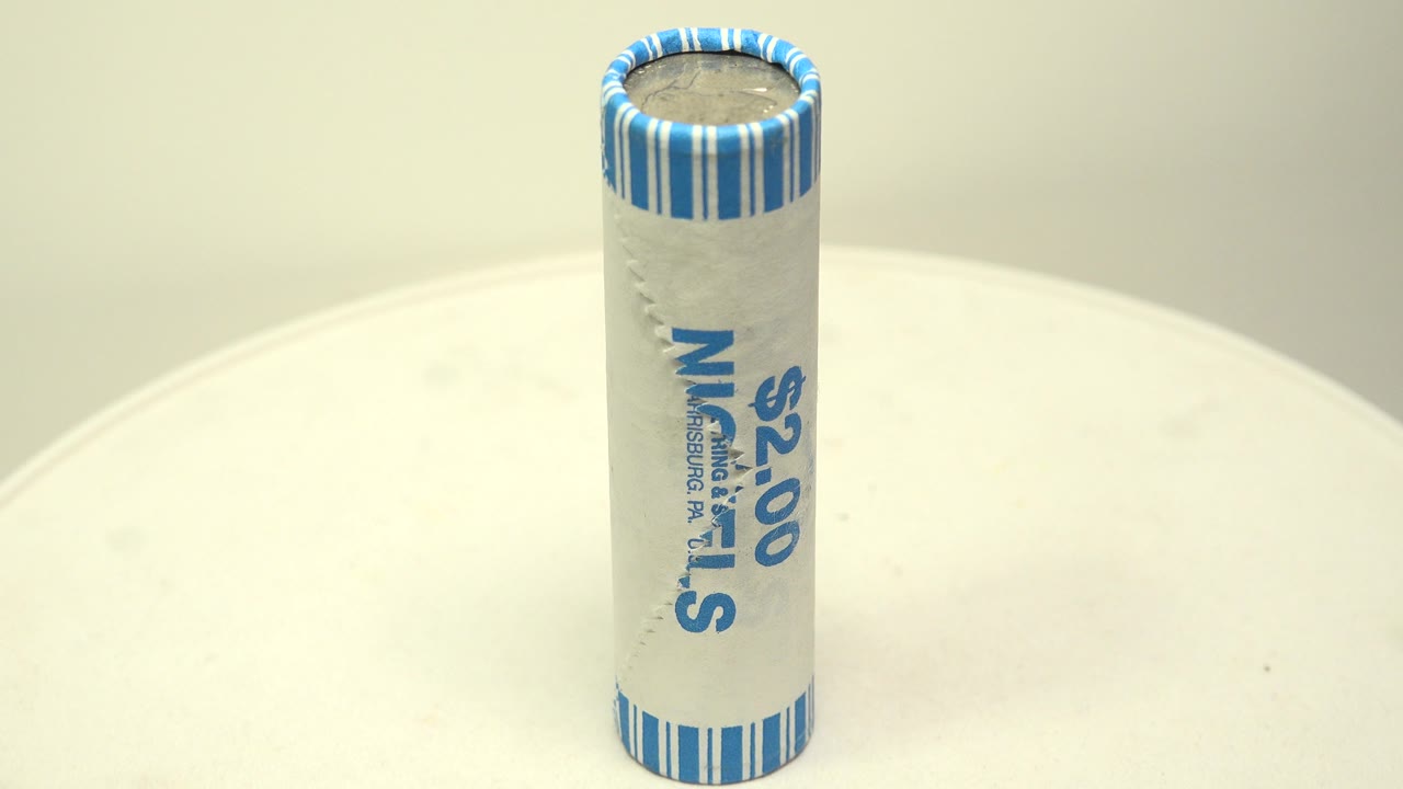 The nickel roll remains upright on the white surface, with the $2.00 marking and blue wrapper text turned more toward the camera, and no visible people.