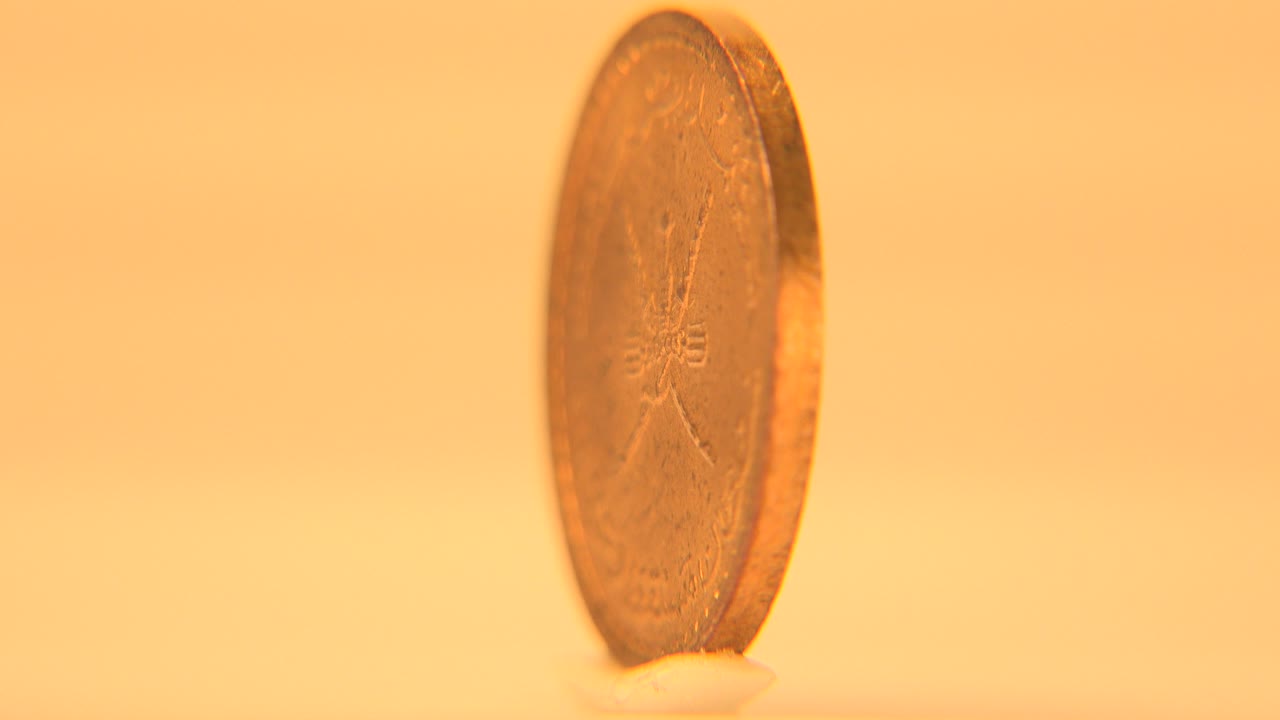 A gold-colored coin stands nearly edge-on near the center of a warm cream-white background, with soft focus and no visible people.