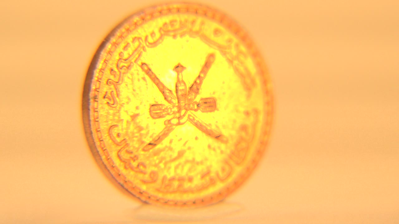 Gold-toned coin shown face-on in soft focus with Arabic script around the edge, crossed emblems in the center, warm amber light, and no visible people.