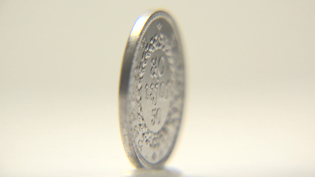The silver coin remains upright near the center of the frame, with a narrow bright rim and blurred engraved details visible against the cream background, no visible people.