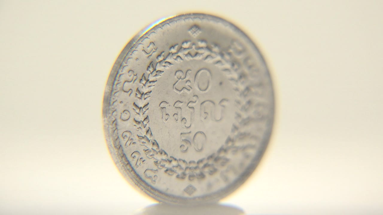 The coin turns toward a fuller front view, showing engraved numerals, decorative wreath-like marks, and curved rim text on a softly lit pale surface, no visible people.