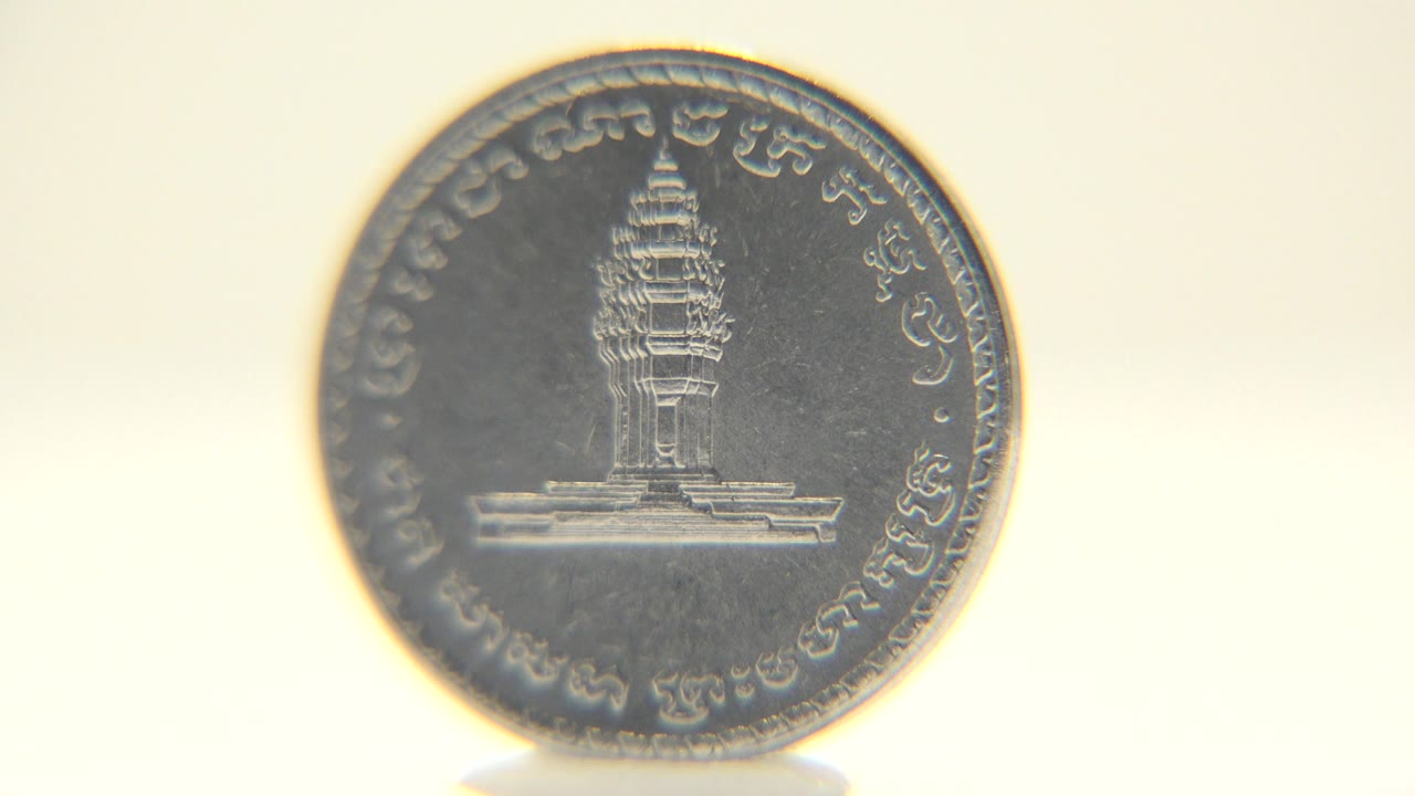 The coin remains front-facing with the building engraving and circular border text visible under soft cream-colored light, with no visible people.