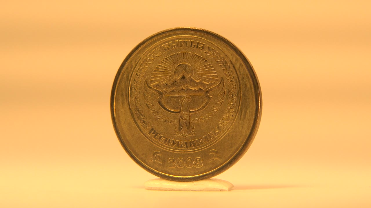 Gold-colored coin shown nearly front-on with an embossed emblem and small text on a warm reflective tabletop, no visible people.