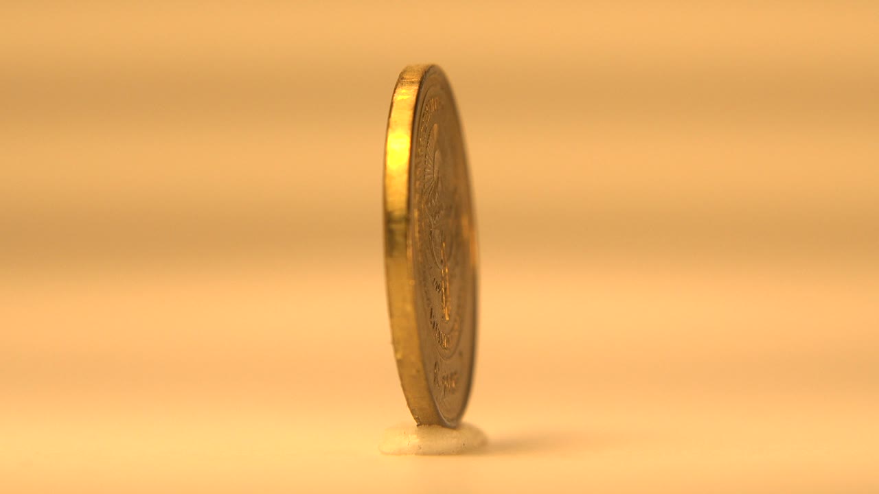 The upright coin remains mostly edge-on, catching a bright highlight along its rim while standing on the small display base, with no visible people.