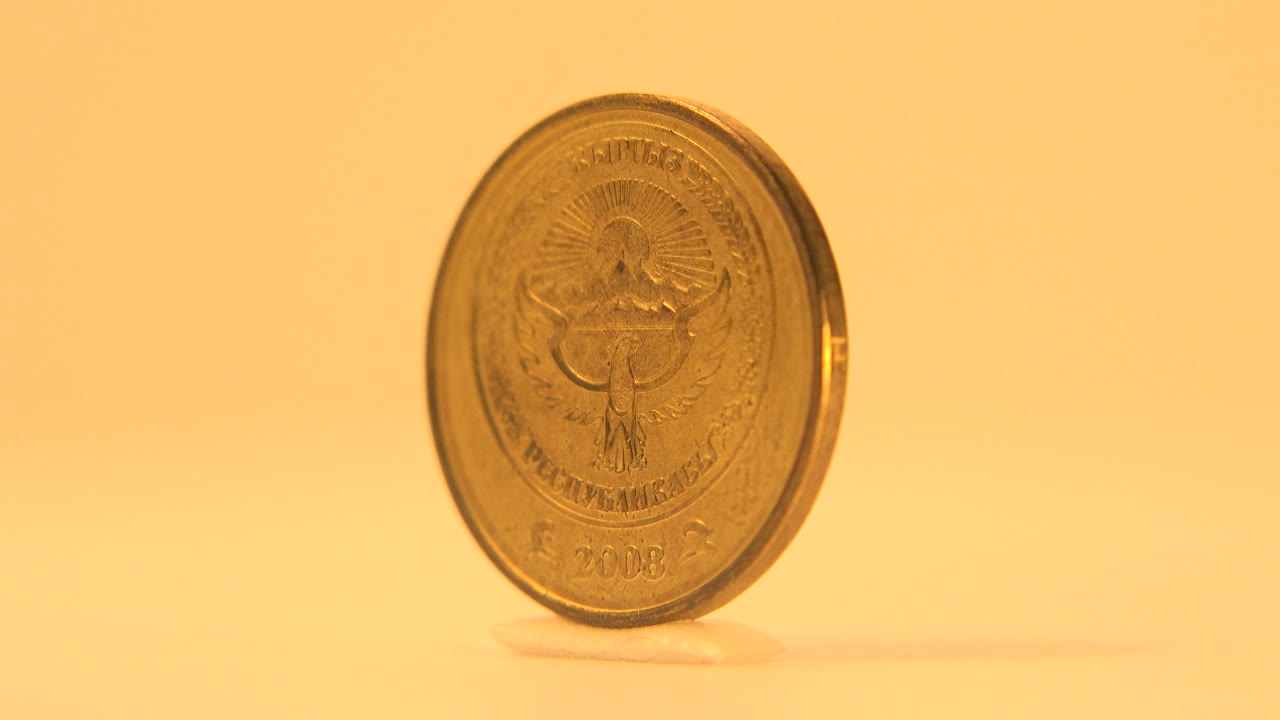 Gold-toned coin showing an emblem side while standing upright on a pale surface against a warm yellow background, with no visible people.