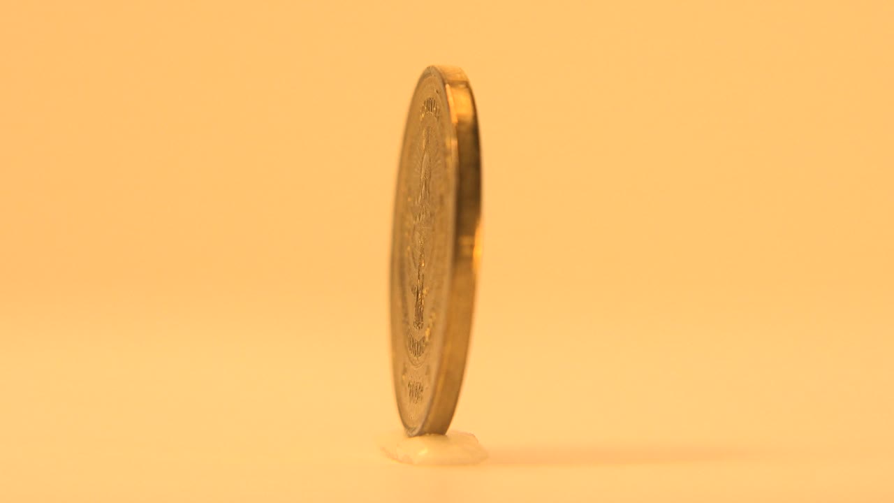 The coin is seen from a narrow edge angle, emphasizing its thickness and reflective rim against the yellow background, with no visible people.