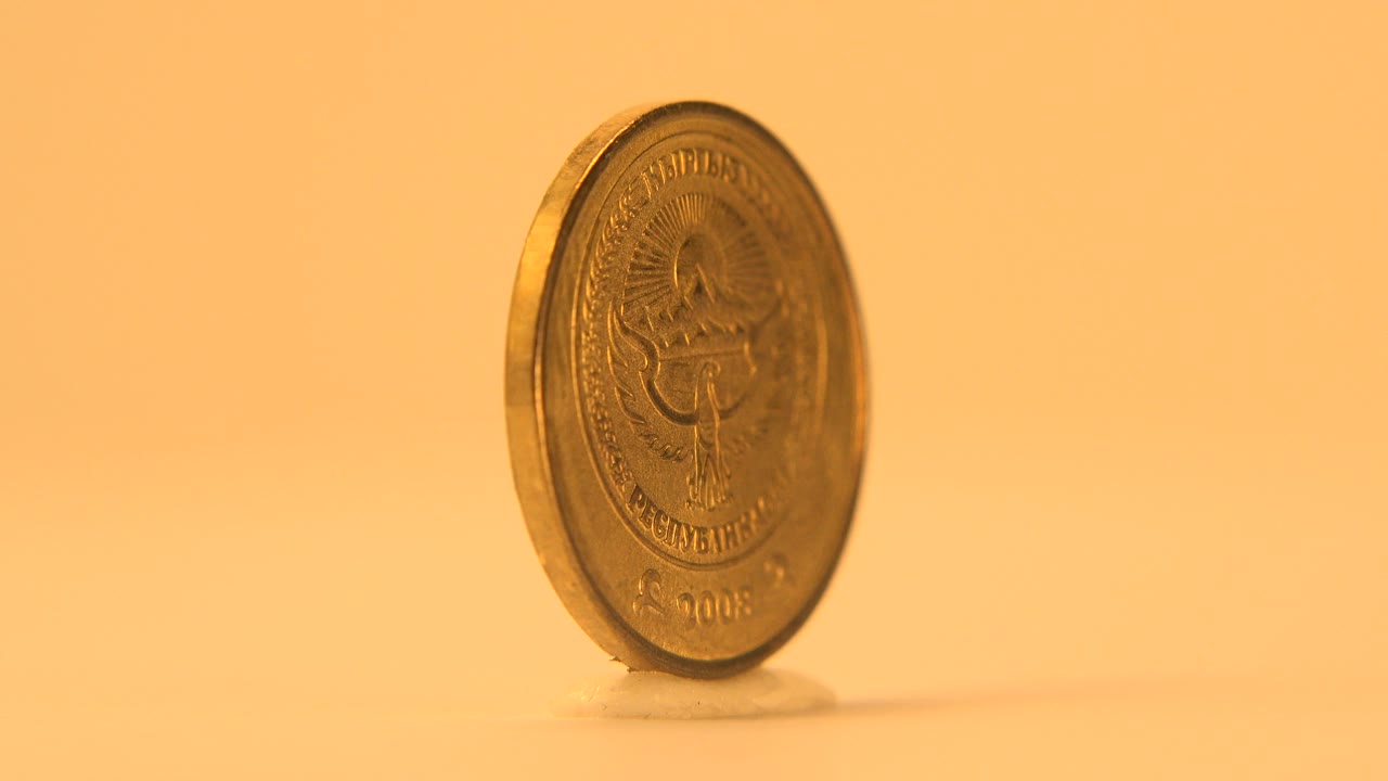 A gold-colored coin stands upright at a left-leaning angle on a glossy warm orange surface, with its reverse side partly visible and no visible people.