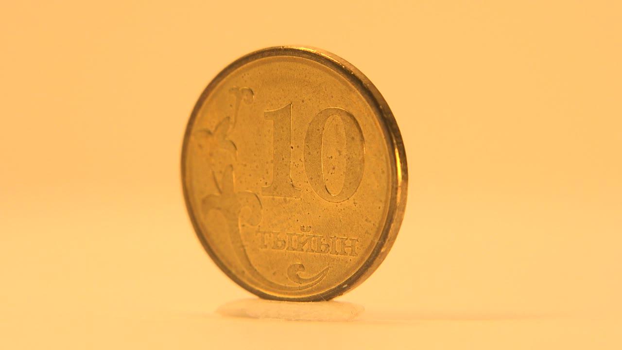 A gold-colored 10-denomination coin stands upright facing the camera on a warm yellow surface, with soft shadowing and no visible people.