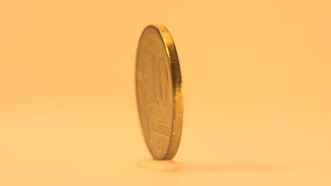 The rotating coin turns almost edge-on, showing its thin rim and a faint face detail against the warm plain background with no visible people.