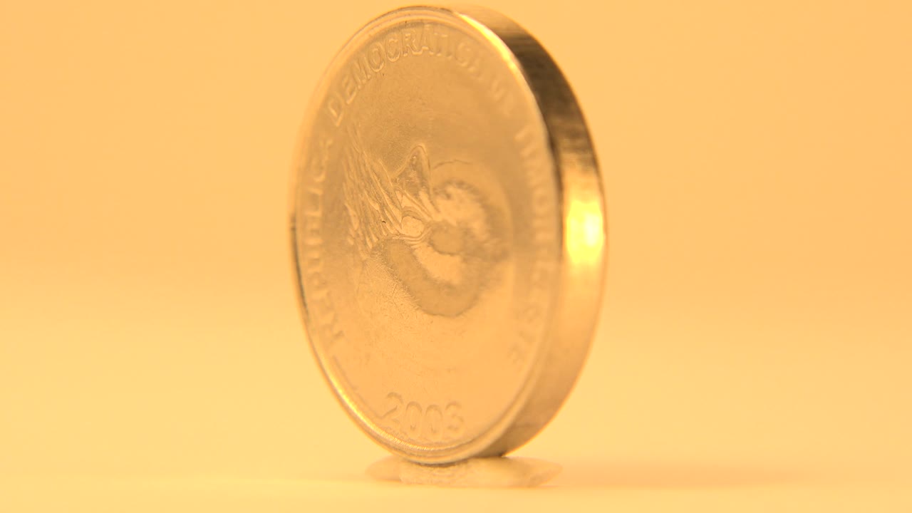 The coin has turned toward a side angle, making the thin edge and bright reflective rim more prominent against the yellow background, with no visible people.