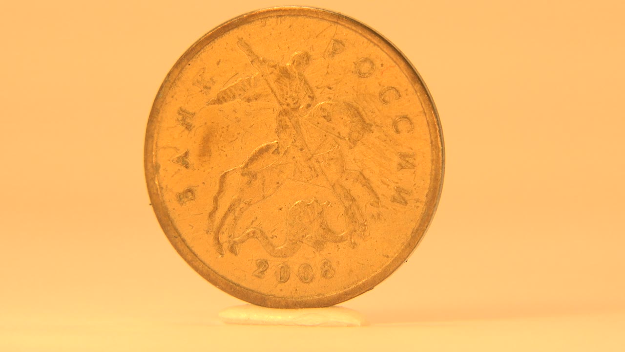 The coin has turned slightly off-axis, making the Saint George face darker and less reflective against the plain orange background, with no visible people.
