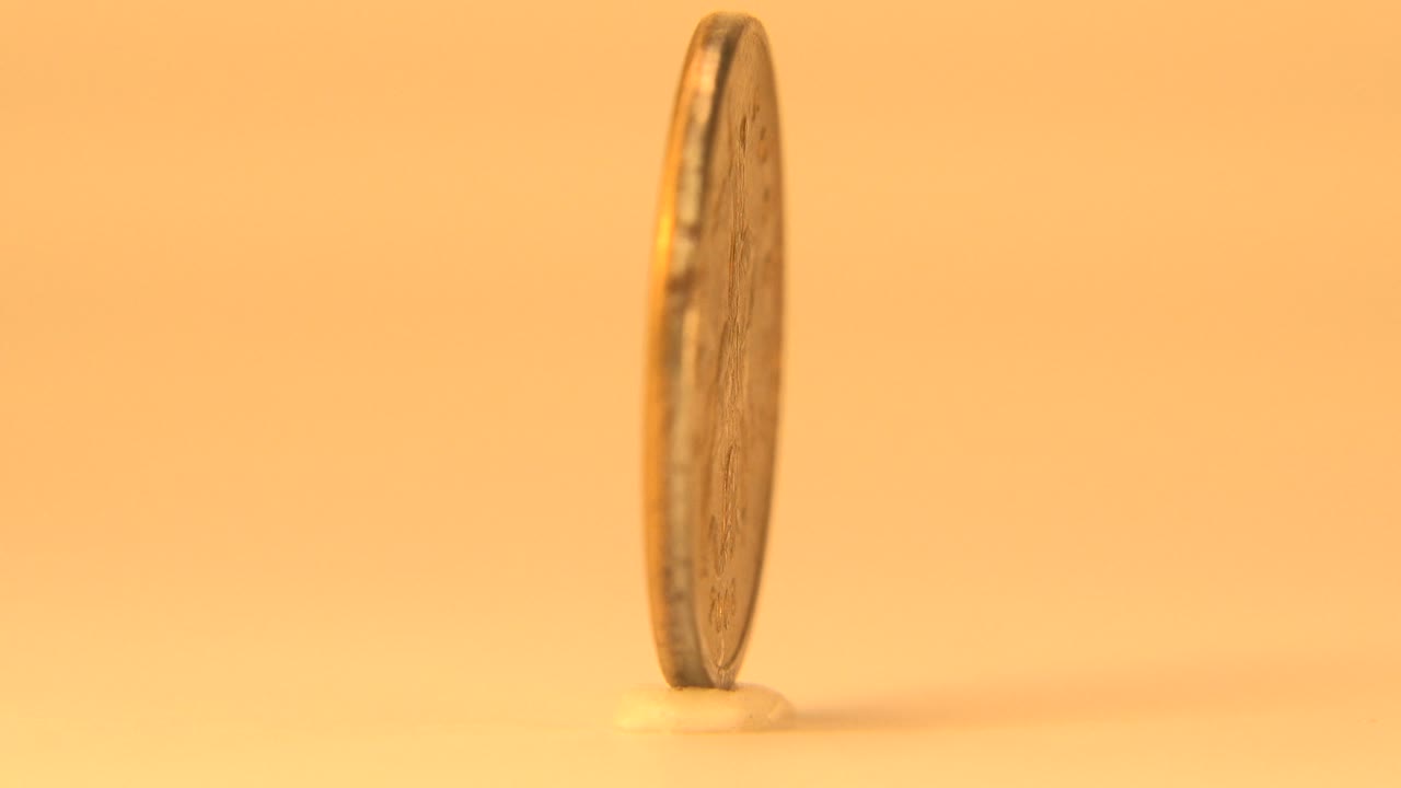 The coin turns to a narrow edge view, showing its thin rim against the warm orange background with no visible people.