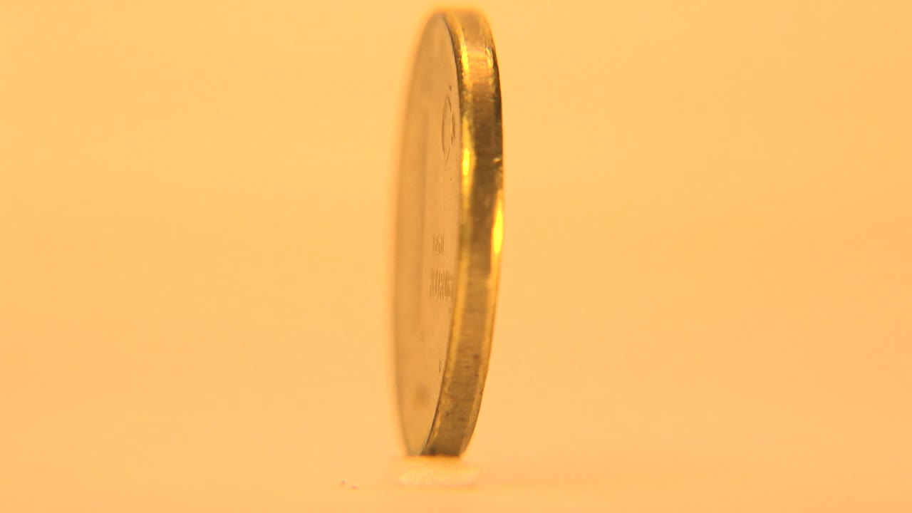 The rotating coin turns to a narrow side profile, showing its thin metal edge against the warm plain background, with no visible people.