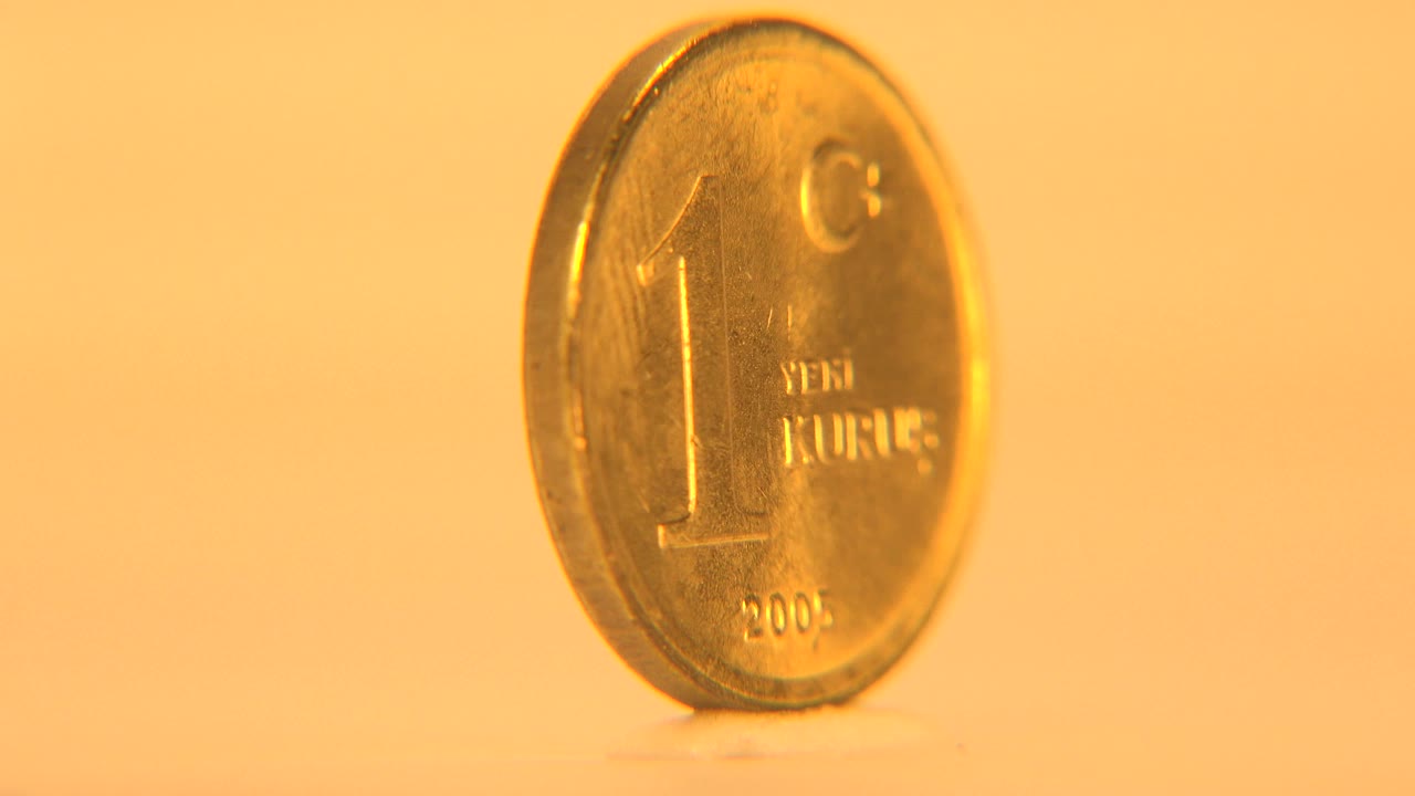 A gold-colored 1 Yeni Kurus coin stands upright at a slight angle on a warm cream background, with shallow focus and no visible people.