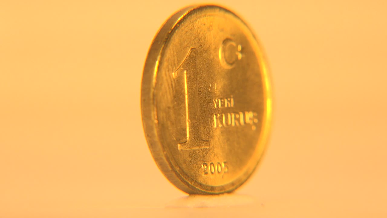 The coin remains upright with its front face partly readable, catching warm highlights along the left rim against the plain cream backdrop, with no visible people.
