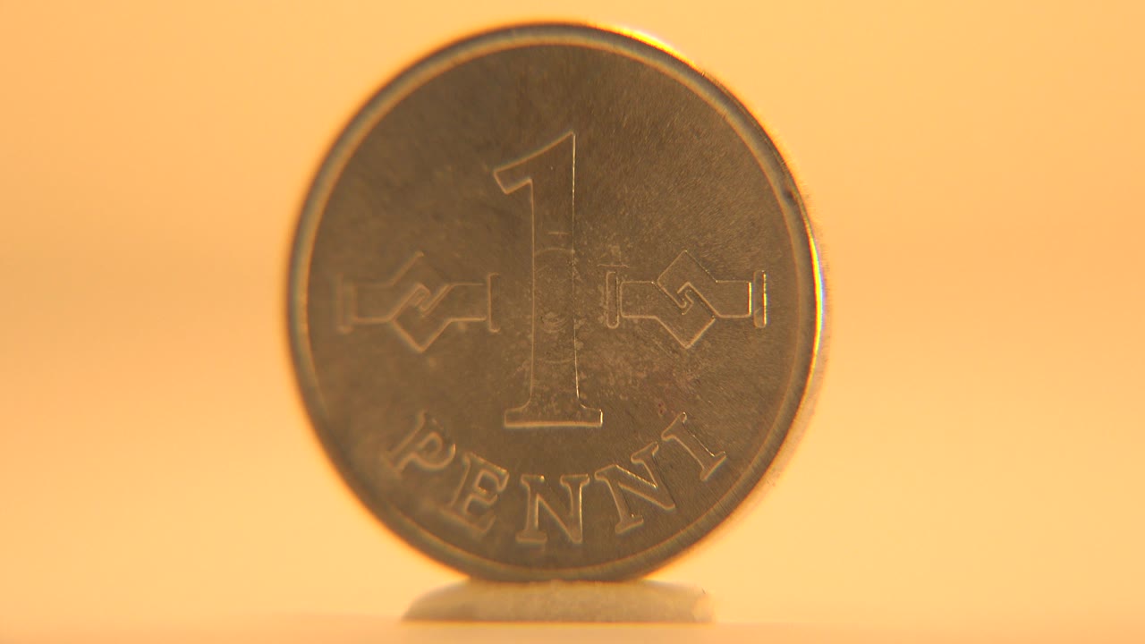 A one penni coin faces the camera while standing upright on a small support against a warm orange background, with no visible people.