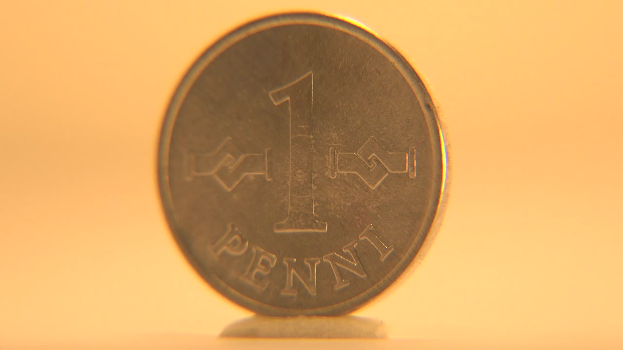 The coin remains nearly face-on with the numeral 1 and PENNI text visible, standing on the pale surface in soft orange light with no visible people.