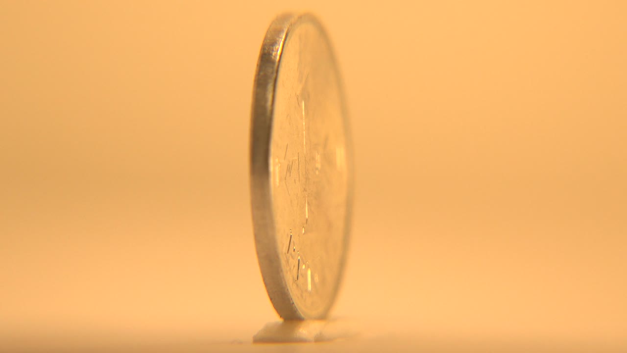 The coin remains mostly edge-on, with a narrow reflective rim and blurred engraved face against the warm tabletop background, no visible people.