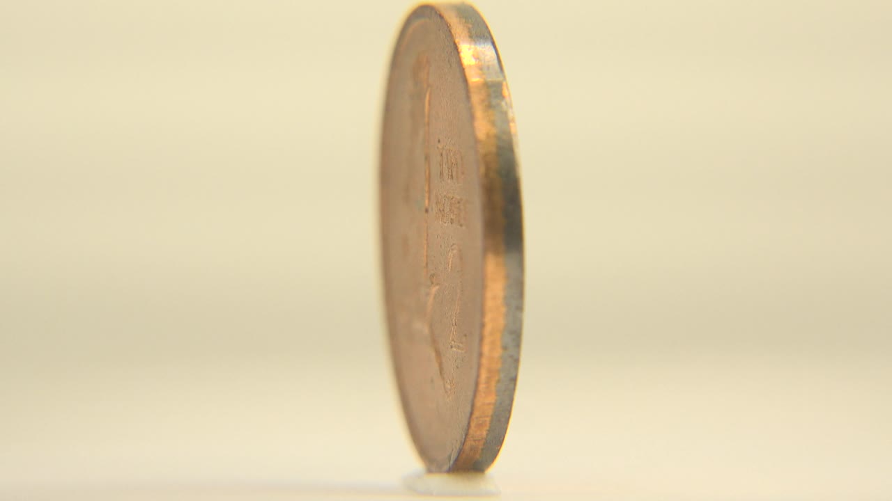 The coin turns to a narrow side profile, showing its rim thickness against the pale background, with no visible people.