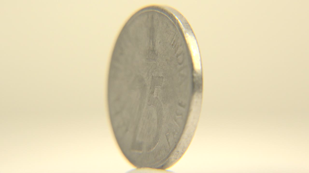 A silver-colored coin stands upright near face-on in soft warm light on a pale surface, with no visible people.