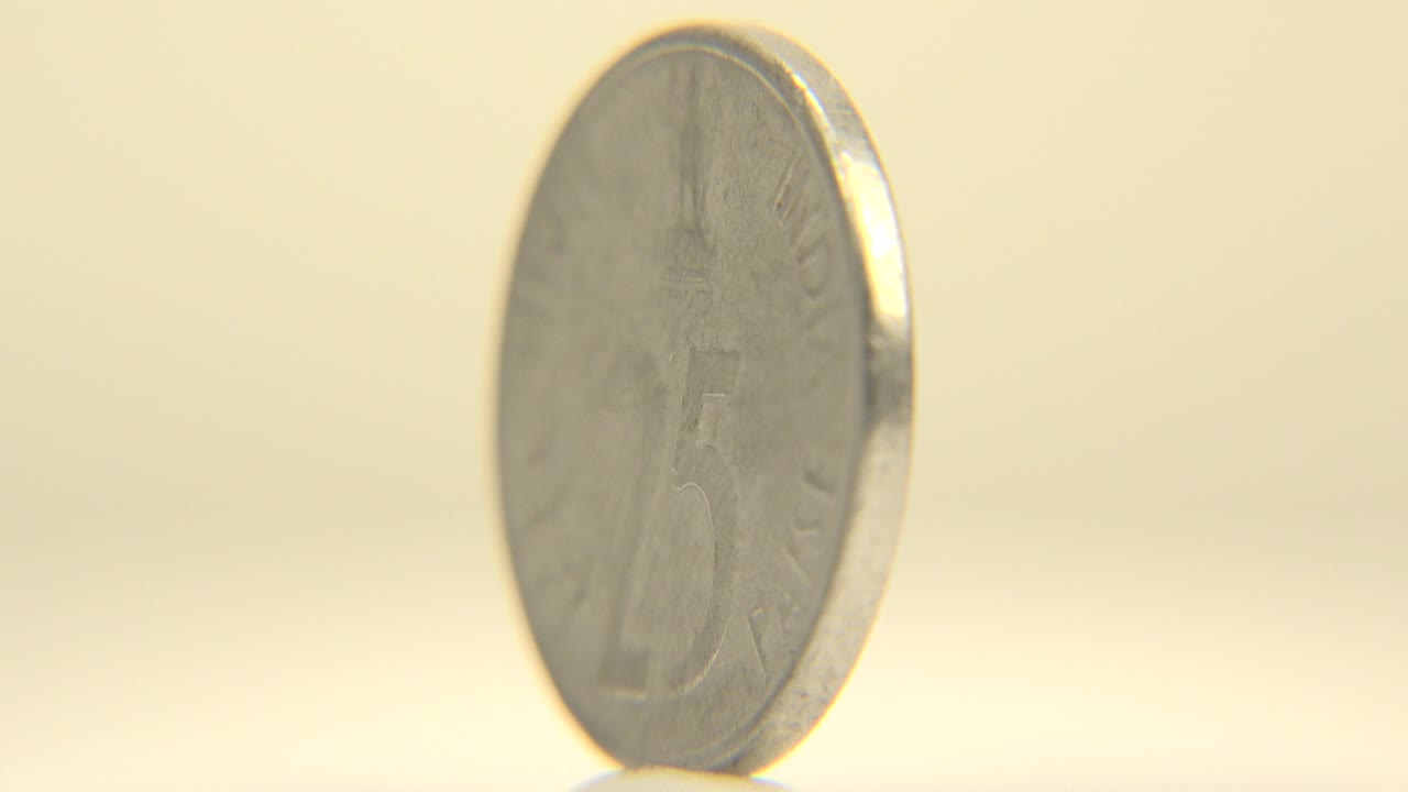 The upright silver-colored coin remains large in frame with a slight turn in its face and a softly blurred pale background, with no visible people.