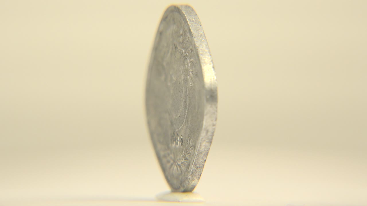 A silver-toned coin balances on its edge in a shallow-focus close-up, with a pale cream background and no visible people.