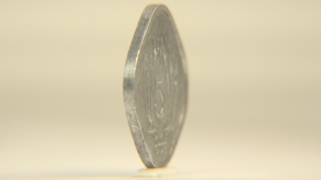A five-value coin rotates partway toward camera, showing a thicker rim and blurred face details on the warm neutral set, with no visible people.