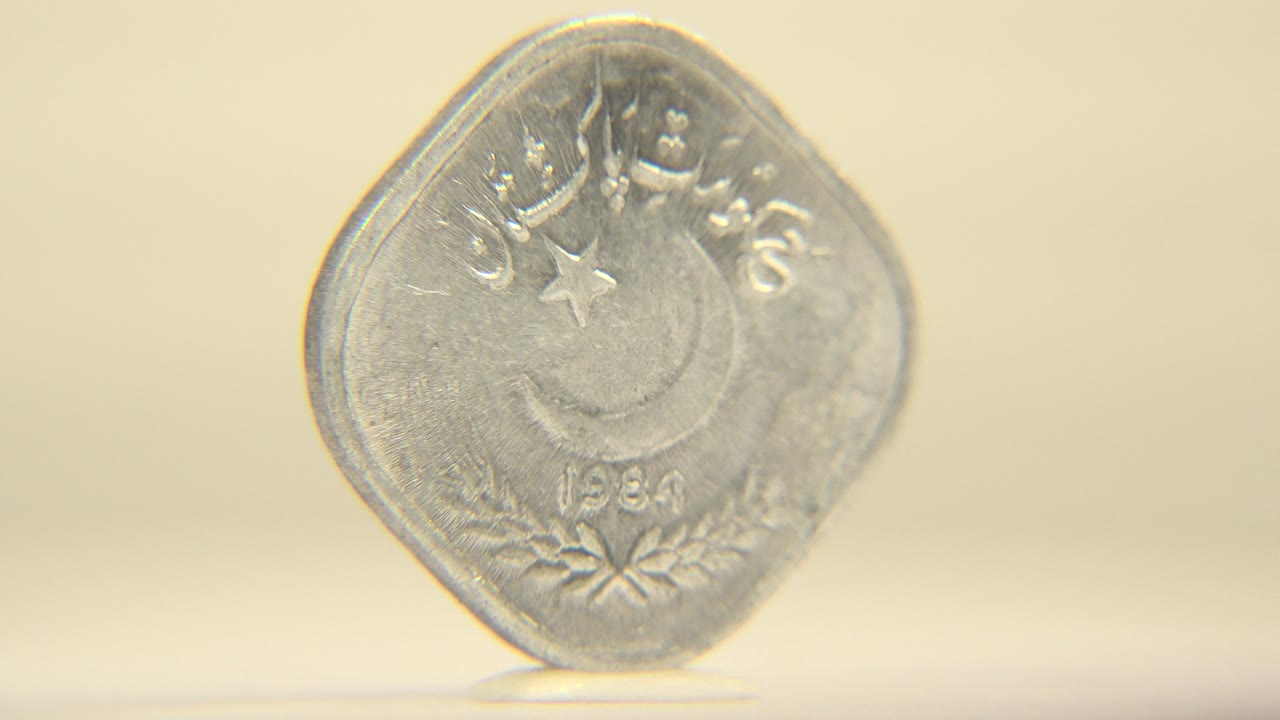 A silver coin stands upright on a cream surface with Arabic and Urdu script, a crescent and star, and the date 1984 visible, with no visible people.